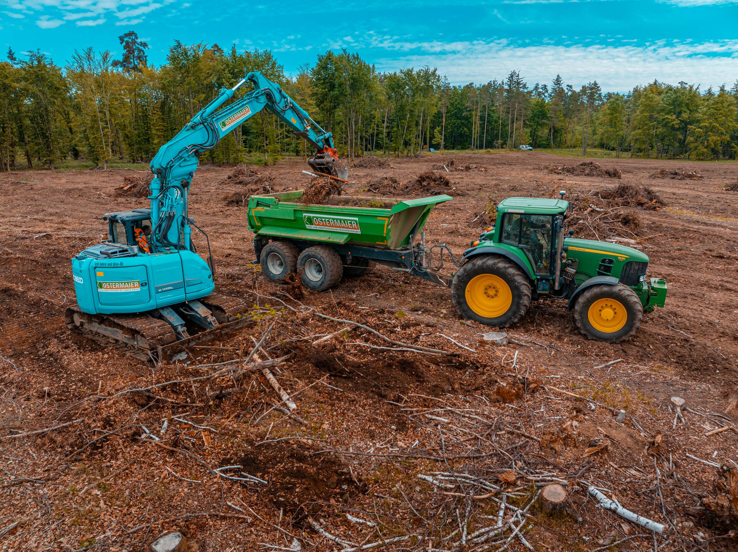 Baufeldrodung von Ostermaier Dienstleistungen: Bagger lädt Wurzelstöcke und Holzreste in Anhänger mit Traktor – professionelle Flächenvorbereitung für Bauprojekte.