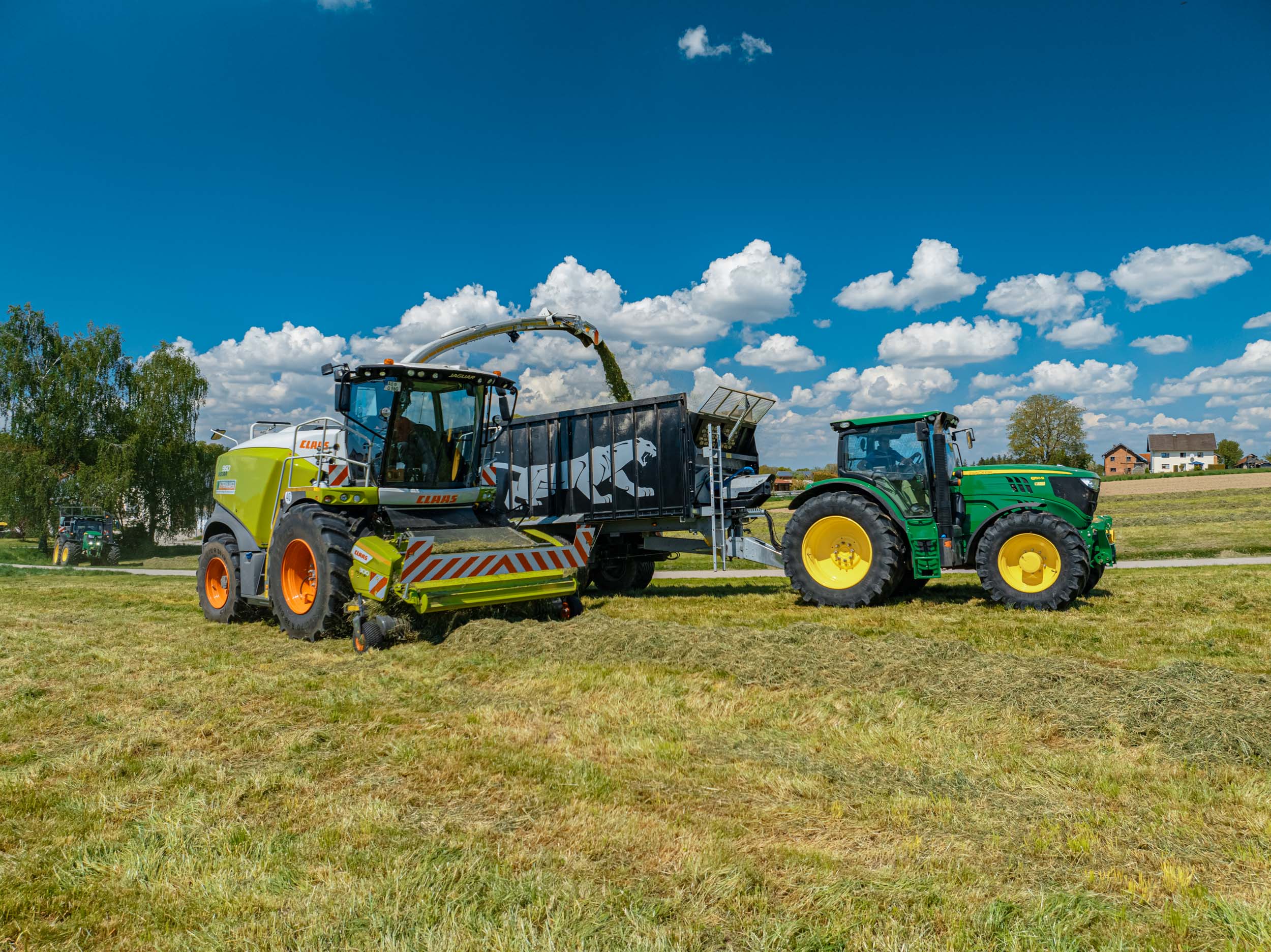 Claas Feldhäcksler übergibt frisch geschnittenes Gras an einen Anhänger, gezogen von einem John Deere Traktor – effiziente Silagearbeiten und landwirtschaftliche Dienstleistungen von Ostermaier.