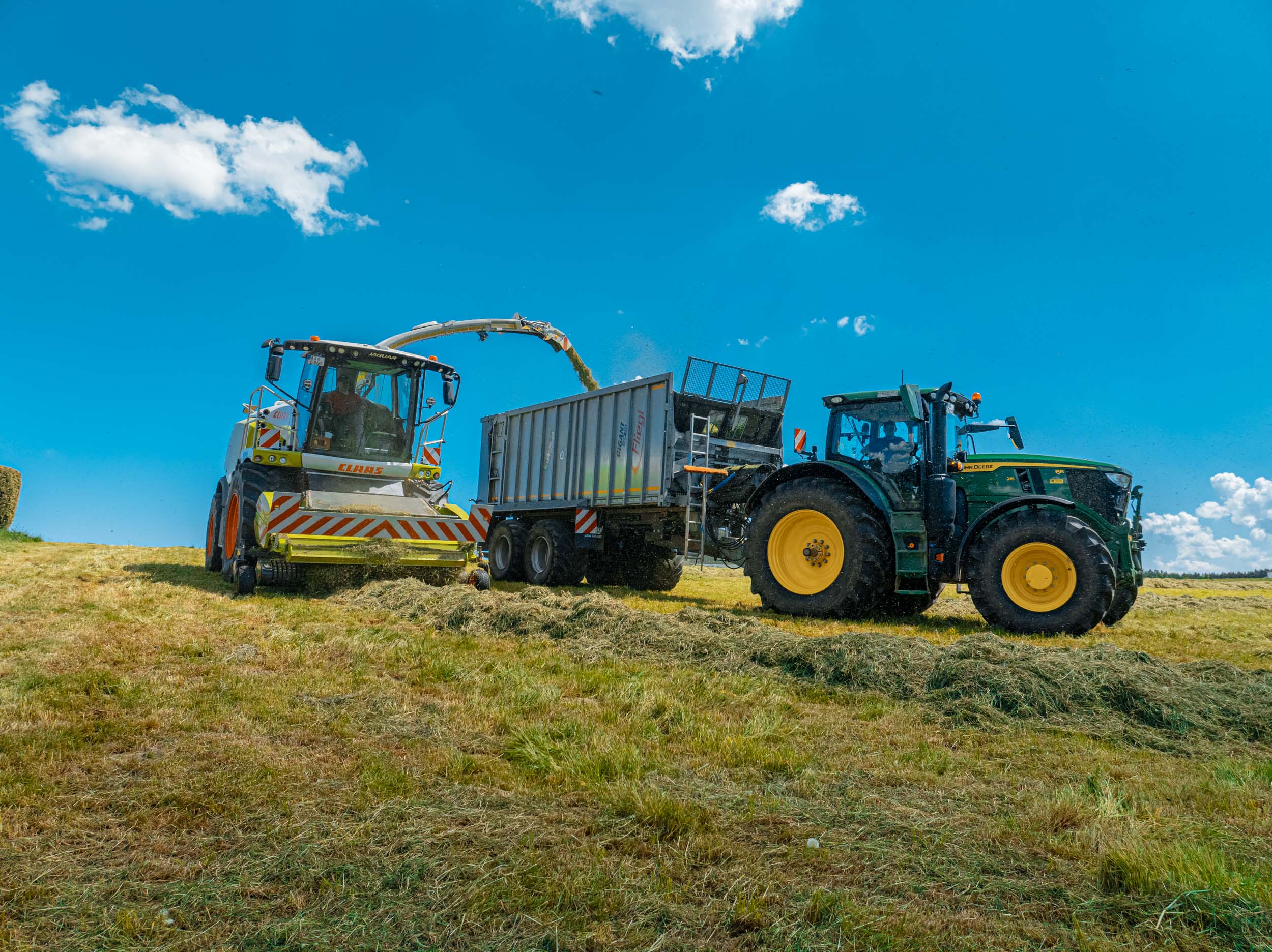 Claas Feldhäcksler übergibt geerntetes Gras an einen Anhänger, gezogen von einem John Deere Traktor – professionelle Silagearbeiten und landwirtschaftliche Dienstleistungen von Ostermaier.