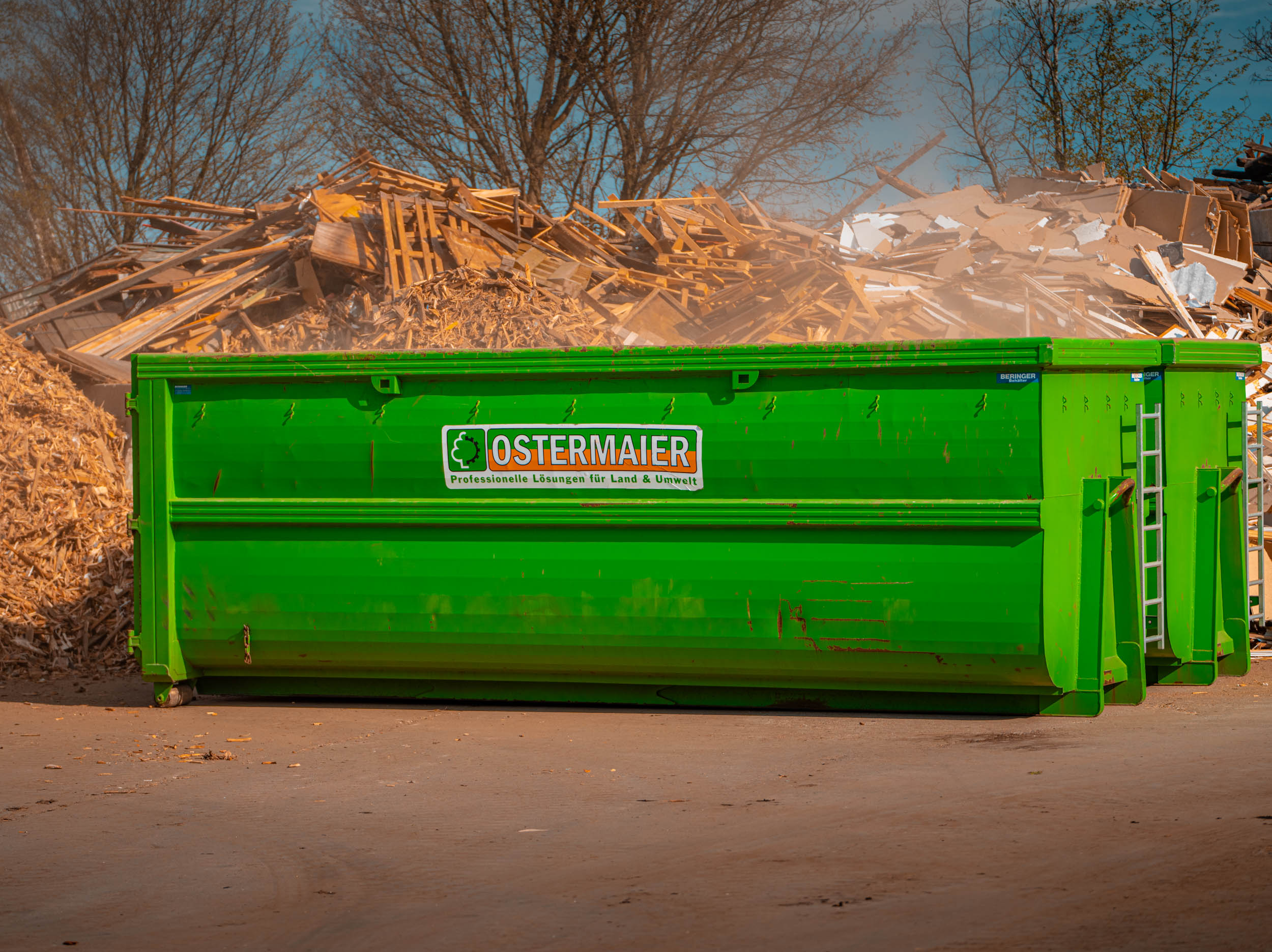 Wertstoffhof bei Ostermaier Dienstleistungen: Grüner Container vor Holzabfällen, professionelle Entsorgung und Recycling für Grün- und Holzreste.