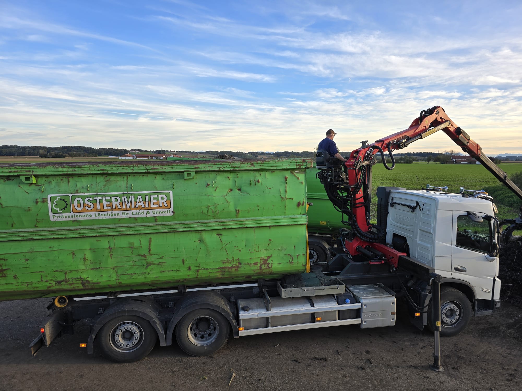 LKW mit Ladekran und großem Container von Ostermaier Dienstleistungen bei der Arbeit auf einem Feld – umwelttechnische Dienstleistungen und Grüngutverwertung.